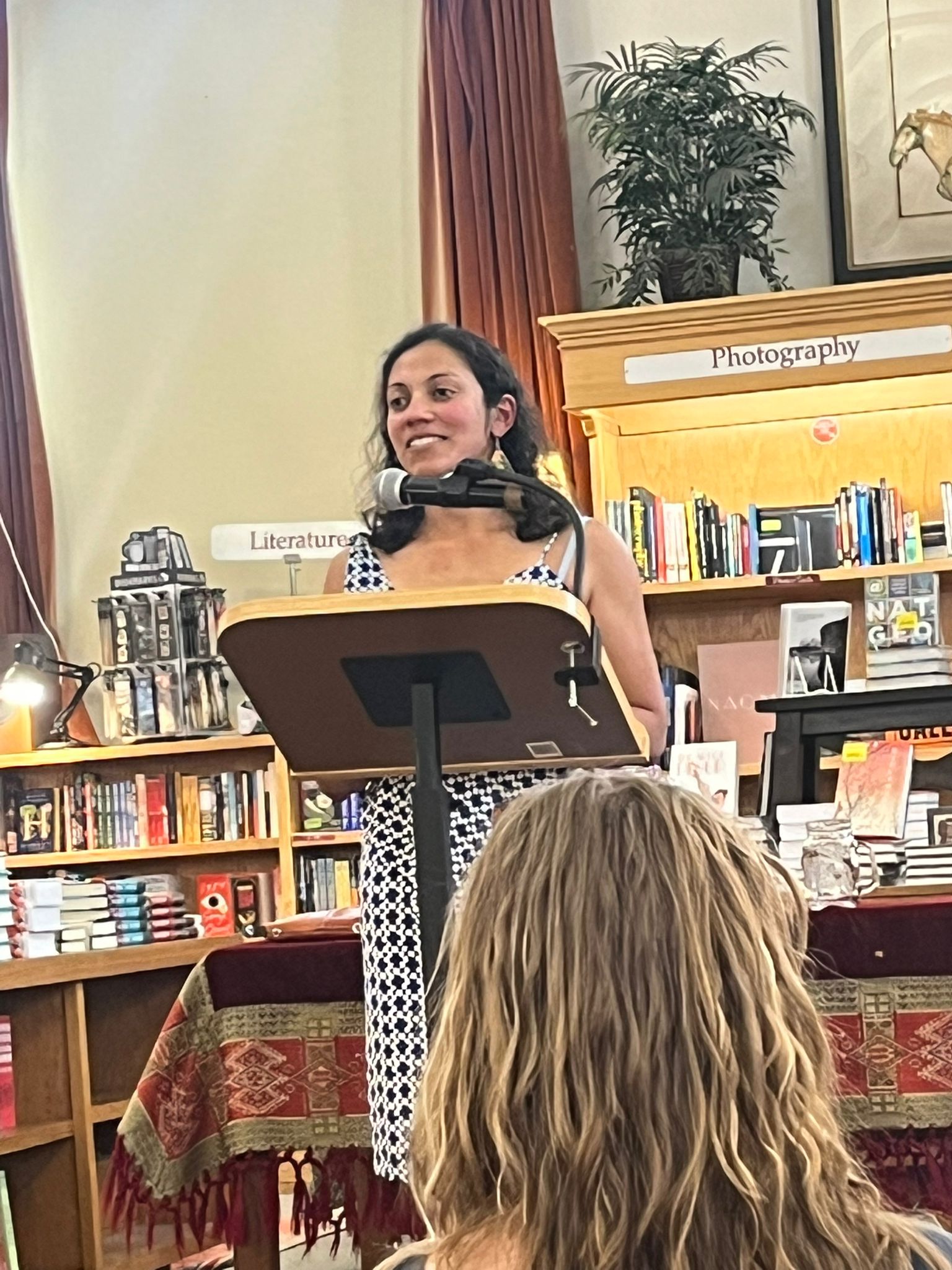 Author Puja Guha speaking at a book signing Events with readers, surrounded by posters and copies of her latest thriller novel.